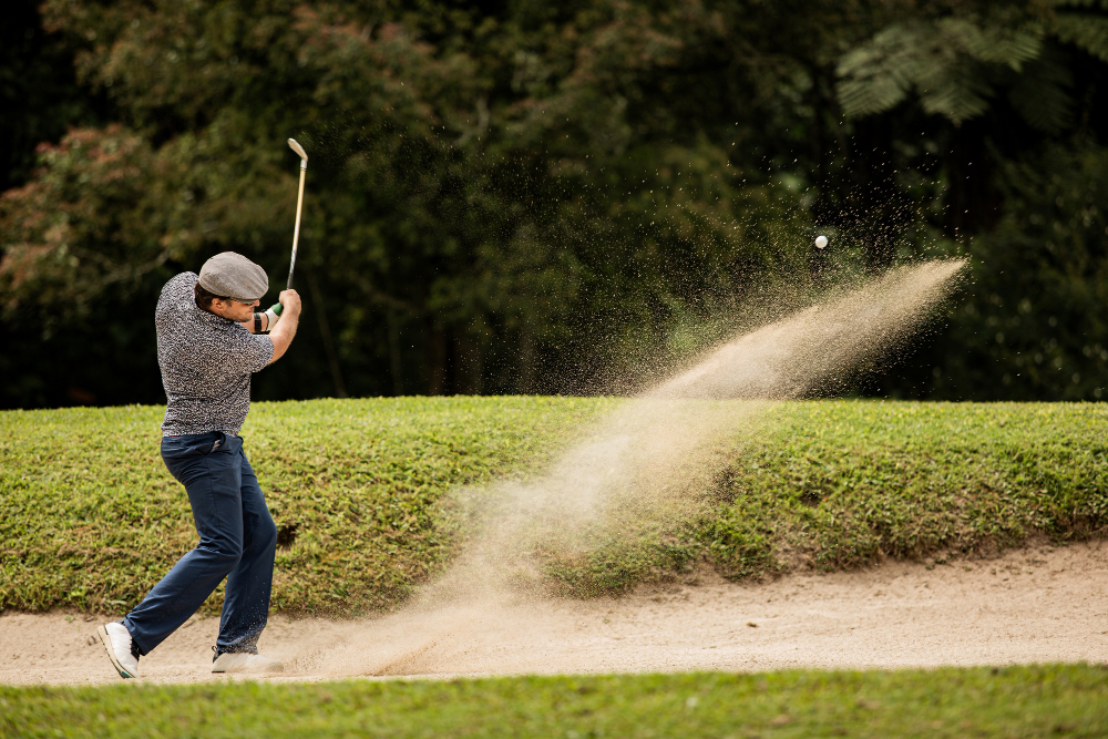 Golfer mid-swing with a golf ball flying out of the sand bunker trailing sand toward the green
