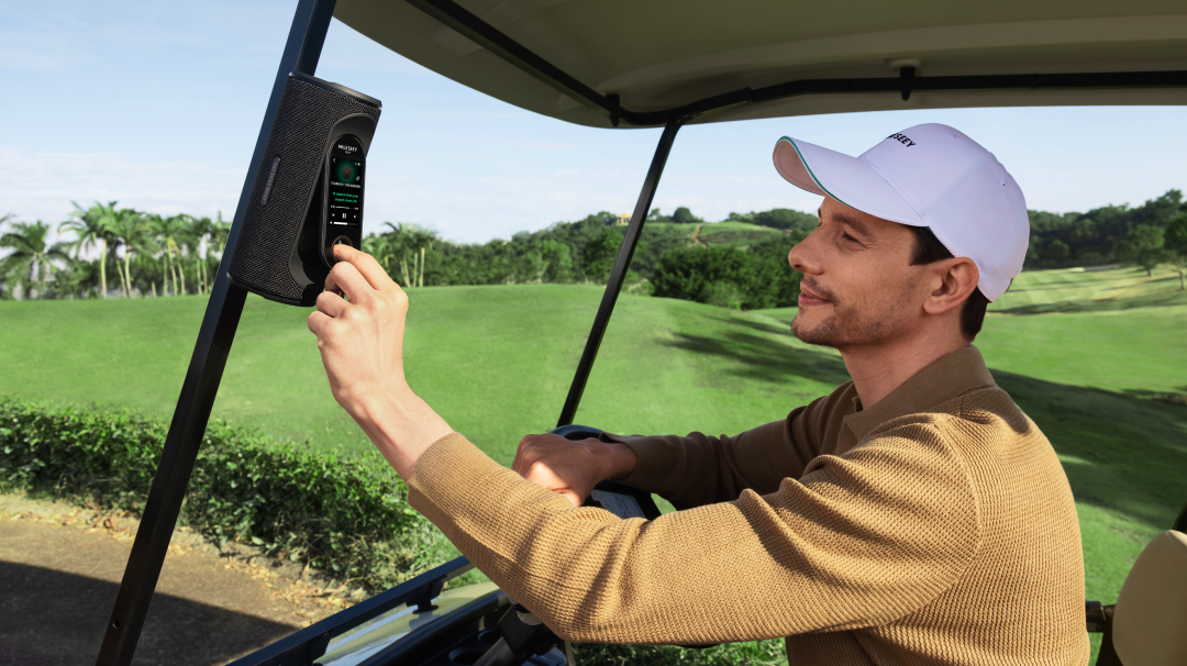 Golfer sitting in a golf cart tapping the MILESEEY GeneSonic Pro Detachable Golf GPS Speaker touchscreen with music playback, magnetically attached to the cart bar
