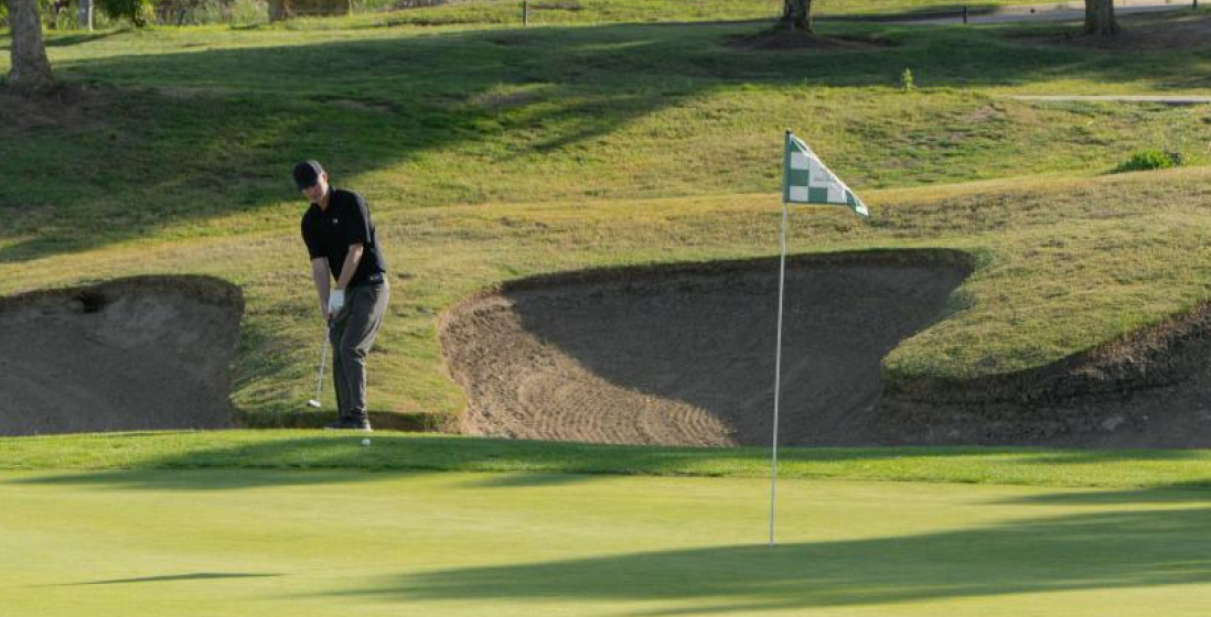 Golfer mid-swing toward a checkered flagstick on a green with a sand bunker behind the pin