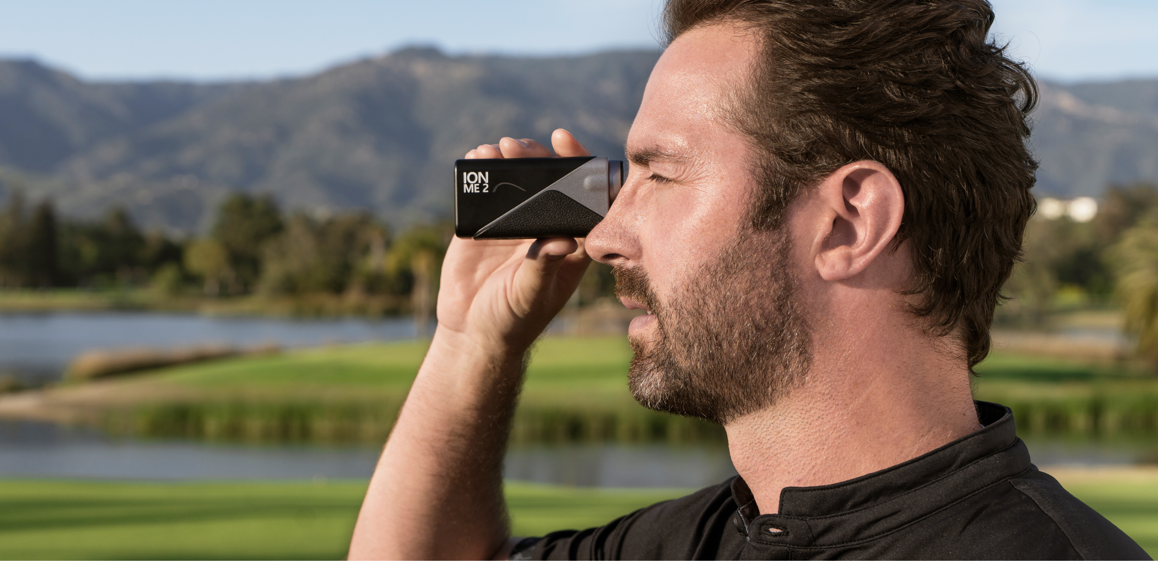 Bearded golfer looking through the Mileseey IONME2 rangefinder aimed at a distant target on a tree-lined course