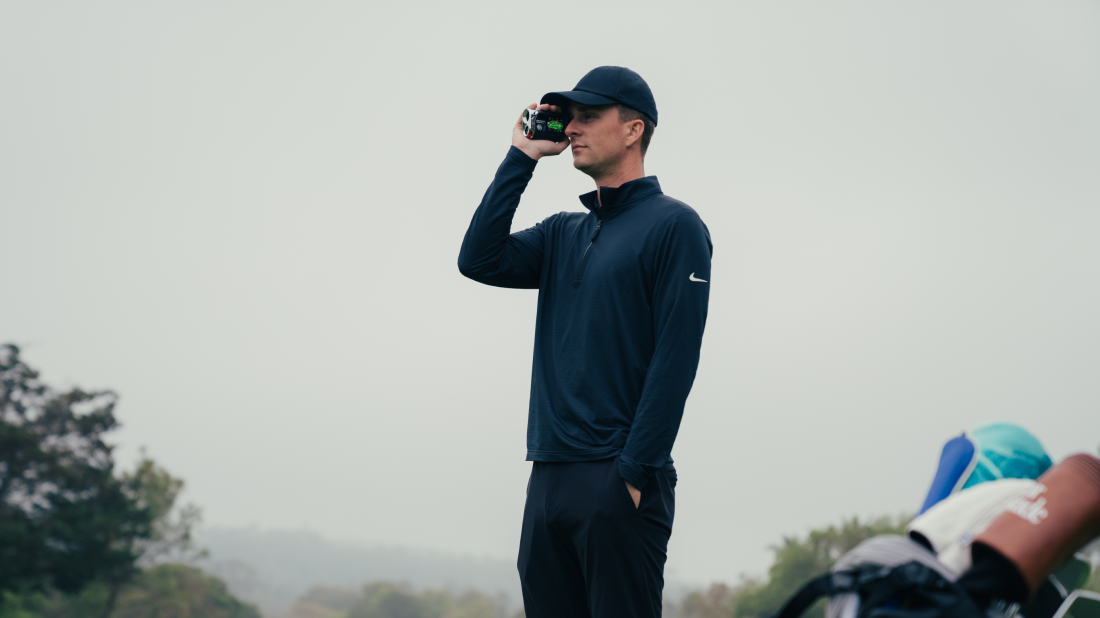 Golfer using the Mileseey GenePro G1 rangefinder on a misty course, golf bag beside him