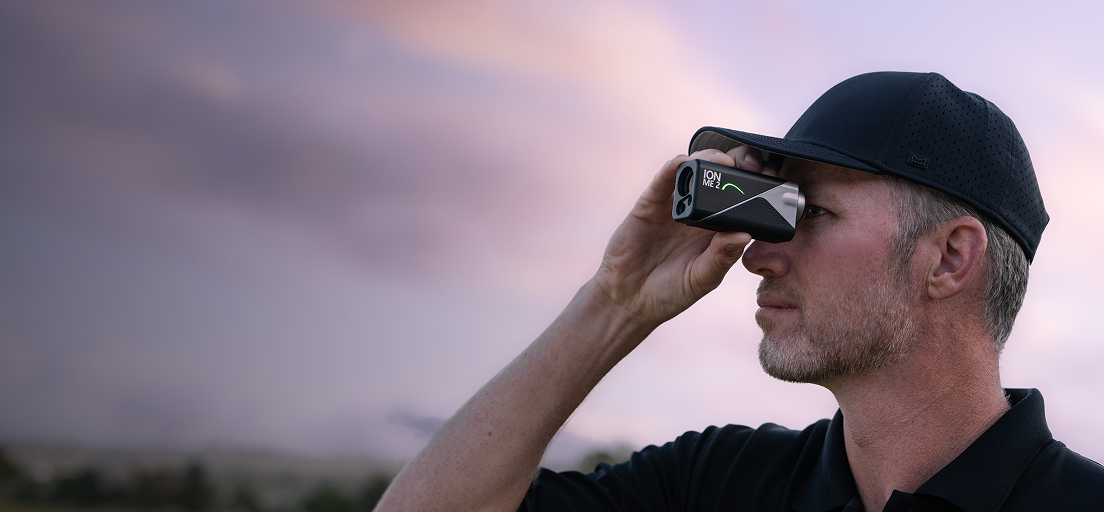 Golfer in a black cap looking through the Mileseey IONME2 rangefinder against a dusk sky