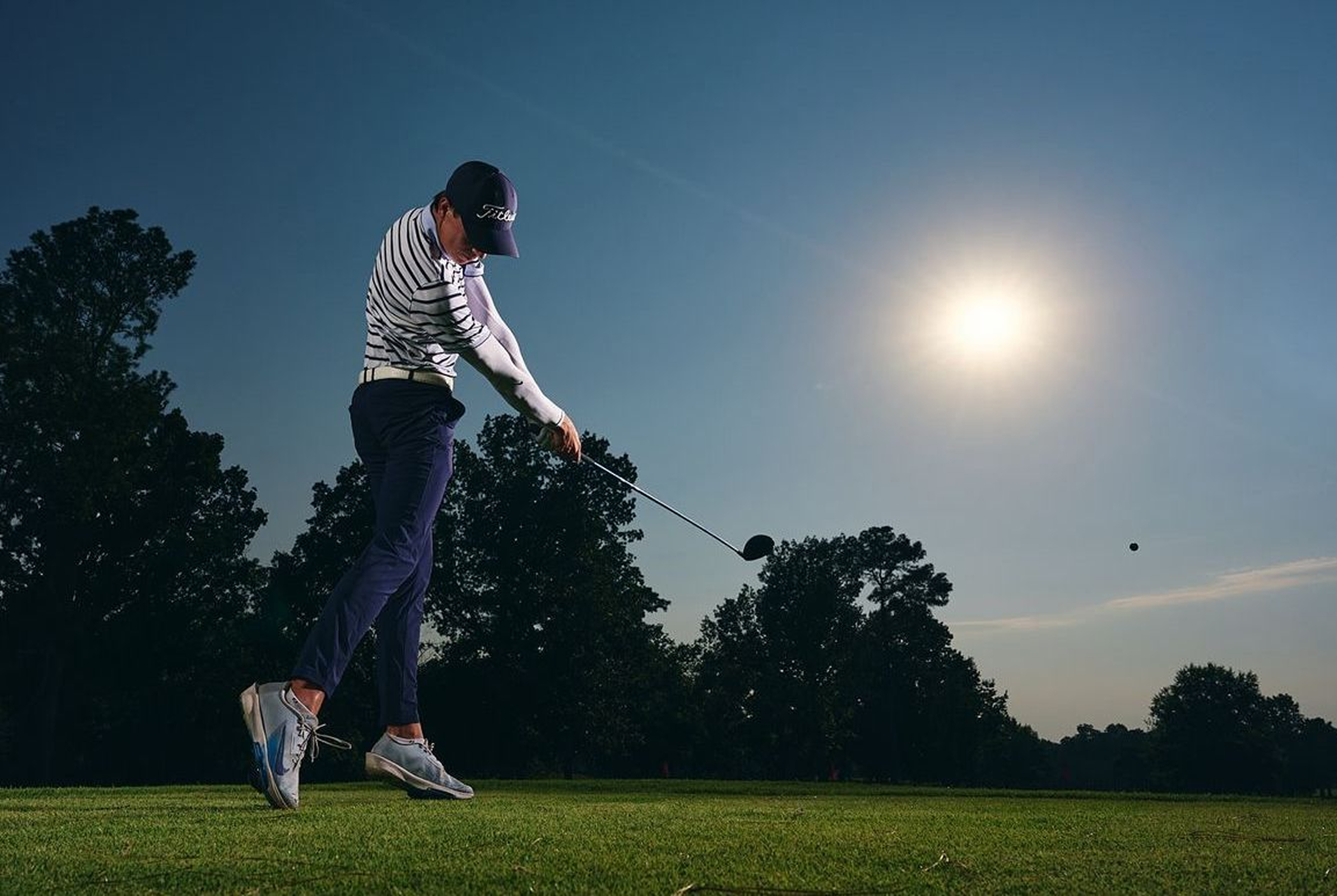 Golfer mid-swing with a driver on a tee box, the ball airborne against a sunlit treeline