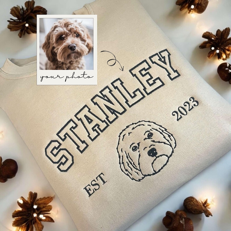 May include: A beige sweatshirt with a black embroidered design. The design includes the word "STANLEY" in a large, bold font, the words "EST" and "2023" in smaller font, and a dog's head. The sweatshirt has a space for a photo of a dog.