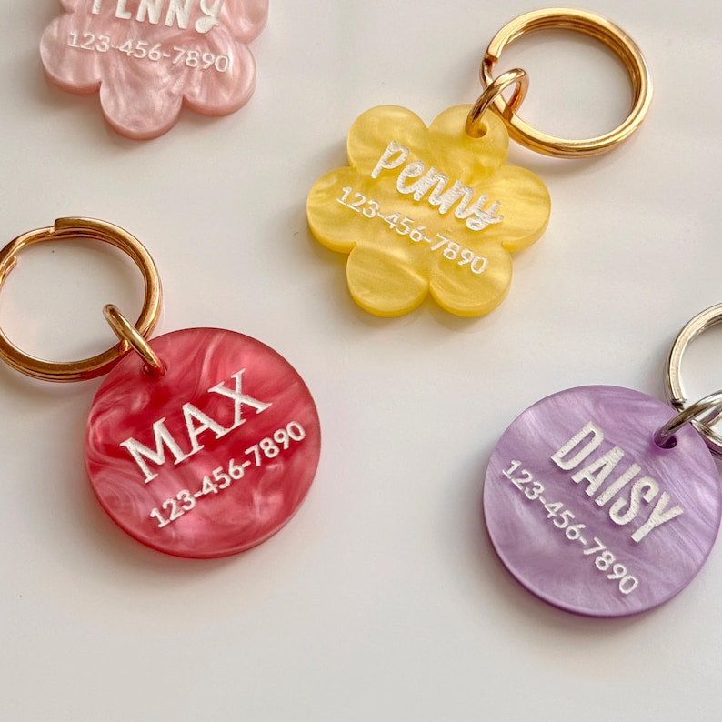 May include: Four acrylic pet tags with gold-toned metal rings. One pink flower-shaped tag reads Penny 123-456-7890. One yellow flower-shaped tag reads Penny 123-456-7890. One red circular tag reads MAX 123-456-7890. One purple circular tag reads DAISY 123-456-7890.