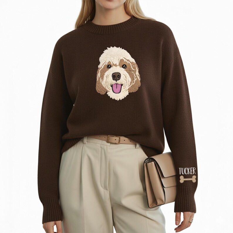 May include: A dark brown sweater featuring an embroidered dog face on the front. The dog has a cream-colored fur and a pink tongue. The sleeve has the name "Tucker" and a bone graphic.