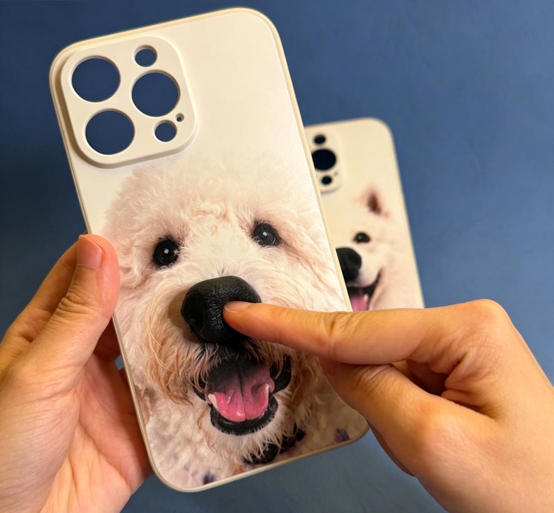 May include: A white smartphone case featuring a high-resolution print of a fluffy white dog with a prominent, raised, three-dimensional black nose. The case includes a cutout for a multi-lens camera system on the top left corner.
