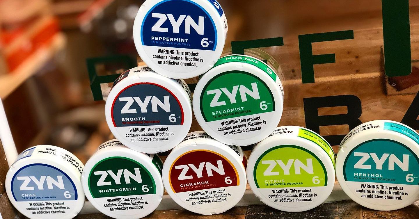 A collection of ZYN nicotine pouches in various flavours laid out on a wooden surface. The 6mg strength tins feature different coloured lids including blue for Peppermint, navy for Smooth, green for Spearmint, dark green for Wintergreen, brown for Cinnamon, lime for Citrus and teal for Menthol. Each tin displays a prominent warning label about nicotine content.