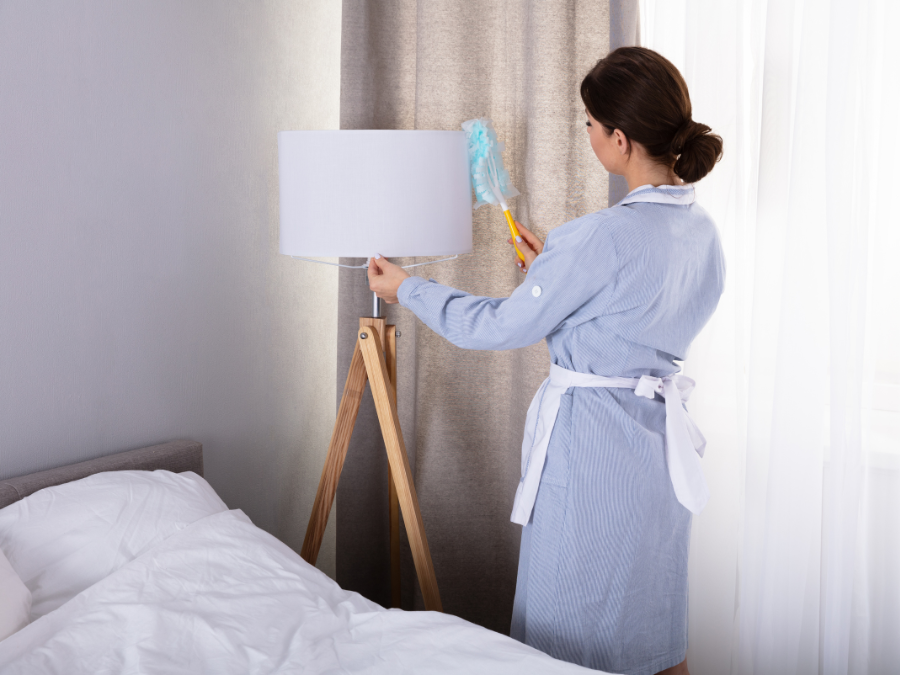Woman dusting white tripod lamp in cozy bedroom.