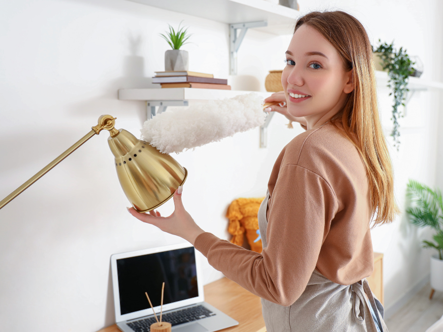 Young woman happily dusting golden lamp in home office.