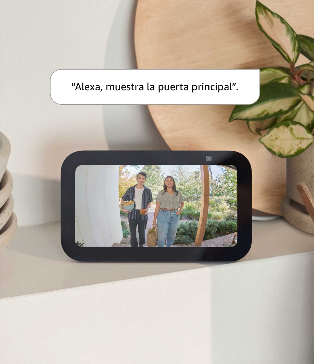 Echo Show 5: Una tablet de 147.3 x 86.4 mm (5.8 x 3.4 in) está en un mueble en posición horizontal. La pantalla muestra a un hombre y una mujer con botanas en un porche frontal. El texto dice “Alexa, muestra la puerta principal”.