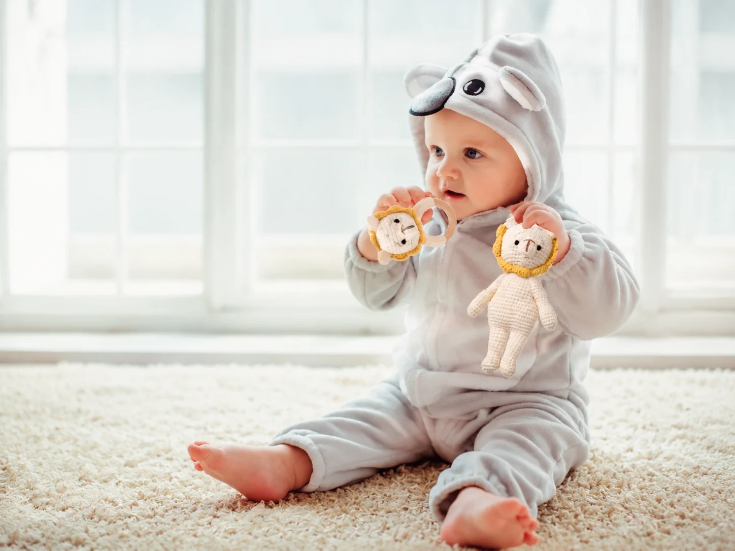 A baby in a koala onesie sitting on a rug, holding a cream crochet lion doll and its matching wooden ring rattle