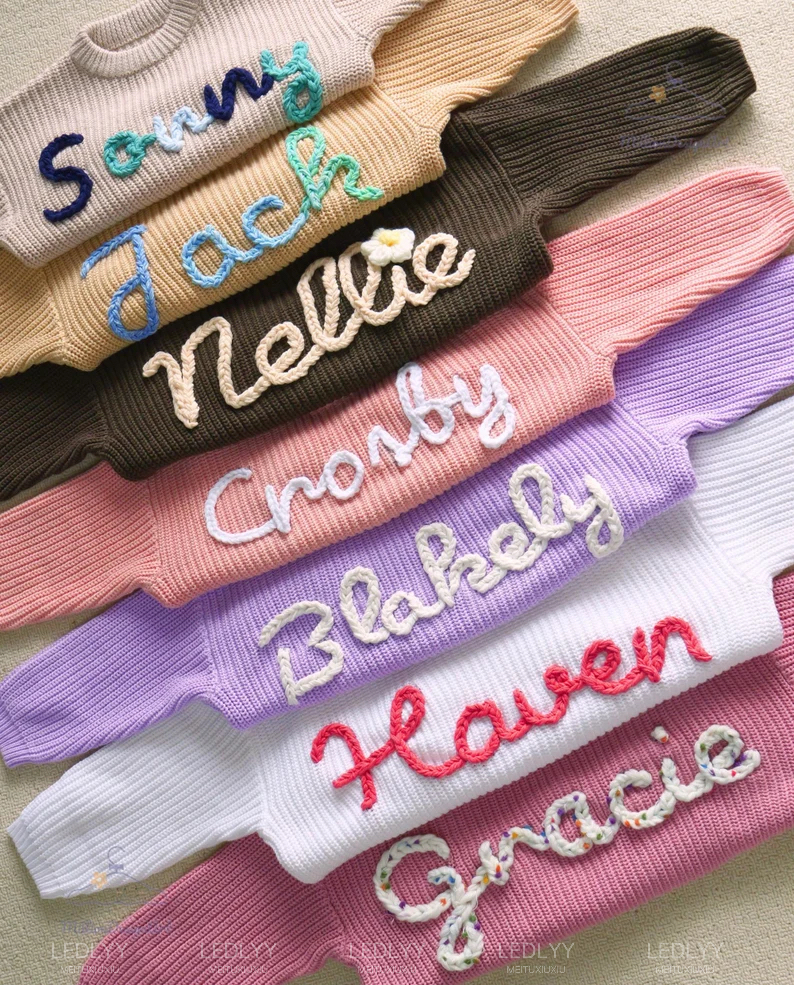 Seven hand-embroidered name sweaters fanned out in different colors, each with a different child's name stitched in cursive