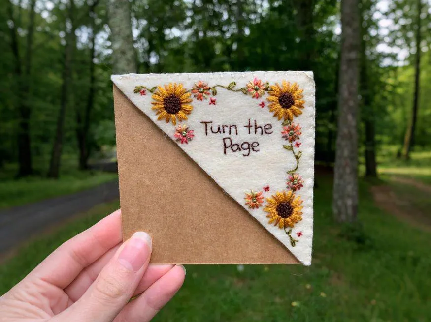 A hand holding up a handstitched felt bookmark embroidered with sunflowers and "Turn the Page", forest path in the background