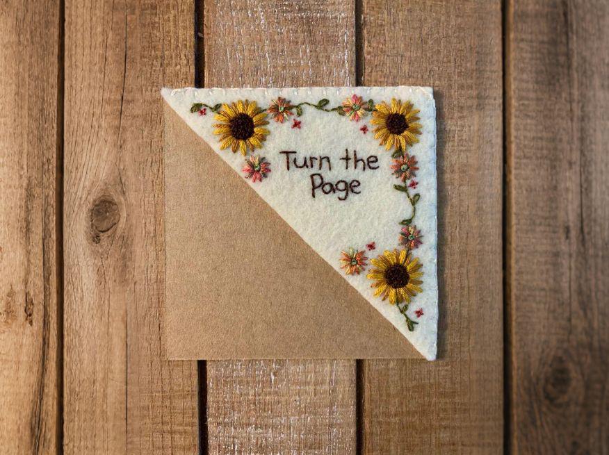 The felt sunflower bookmark laid flat on a rustic wooden surface, showing the embroidery detail and kraft-colored backing
