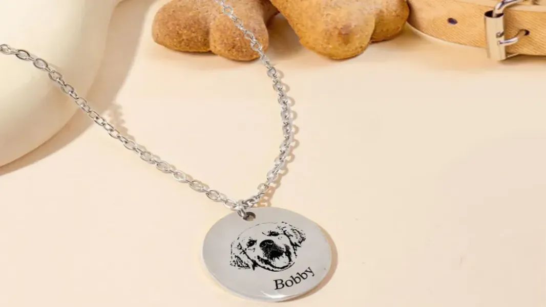 Close-up of a woman's hand holding a small engraved pendant necklace with a laser-etched pet photo, soft afternoon window light