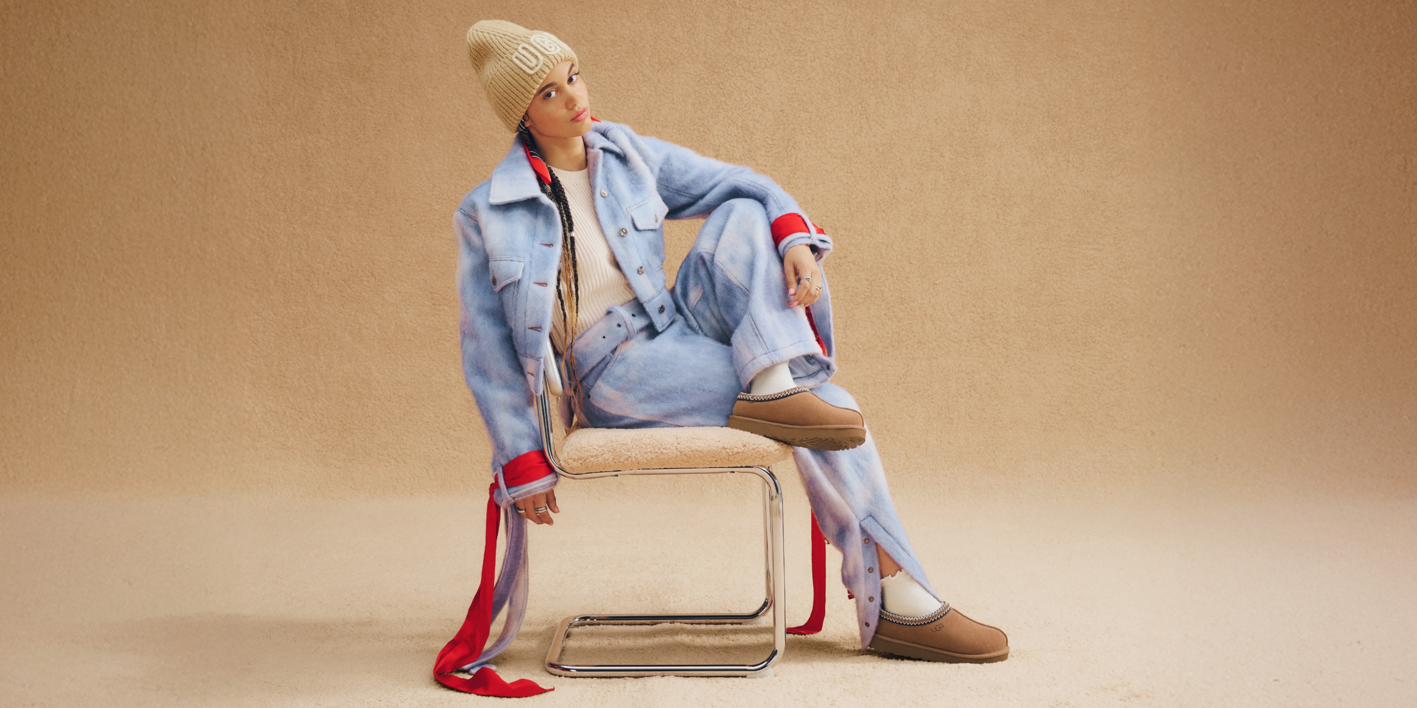 A person sitting sideways on a chair, against a beige backdrop wearing UGG Tasman II, light blue tie-dye denim jacket and pants, a cream turtleneck sweater, an UGG beanie, and a red scarf draped down to the floor.