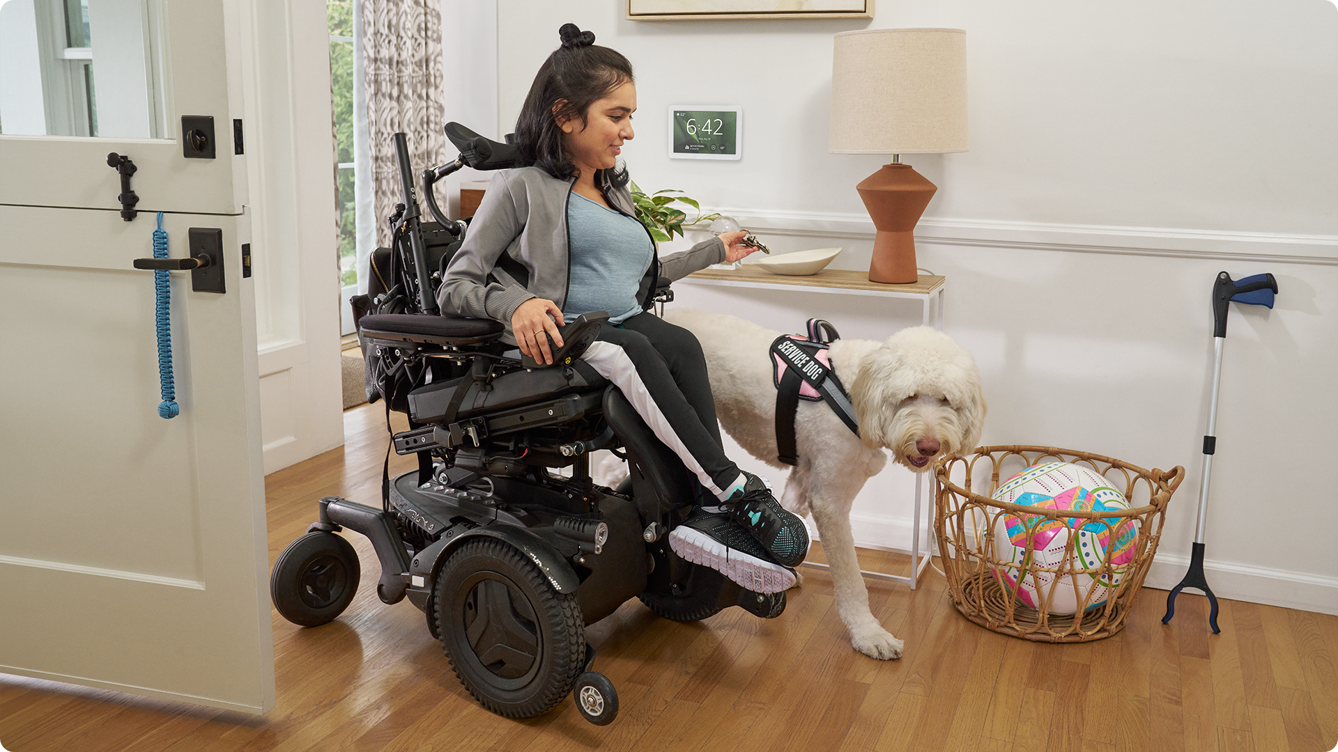 In an entryway, a woman in a motorized wheelchair with a service dog drops her keys in a bowl. Next to her on the wall is an Echo Hub showing the time: 6:42 pm. 