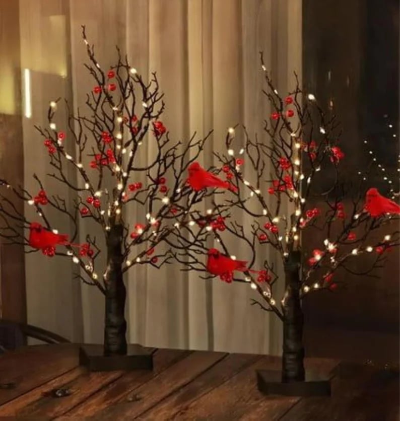 May include: Two decorative black tree sculptures with red cardinal bird ornaments, red berry clusters, and warm white LED lights. The trees are set on a wooden table against a neutral background.