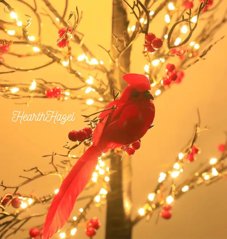 May include: A vibrant red cardinal perched on a branch of a decorative tree. The tree is adorned with small, warm white lights and red berries. The image has the text "HearthHazel" in the lower left corner.