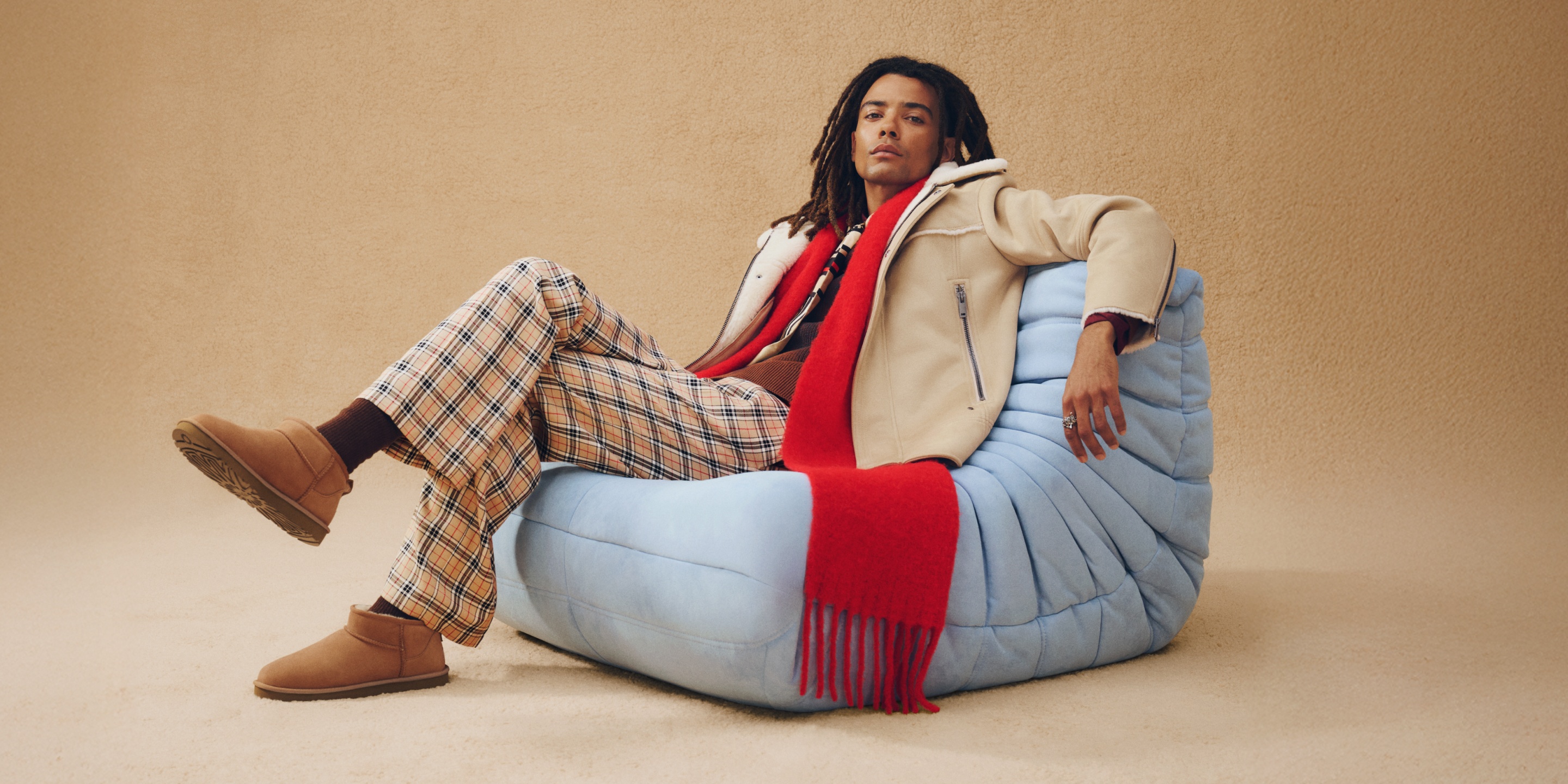 Una persona adagiata su una poltrona imbottita azzurra davanti a uno sfondo beige indossa stivali UGG Classic Ultra Mini da uomo in tonalit&agrave; Chestnut, calzini marroni, pantaloni con motivo plaid, giacca di sherpa in tonalit&agrave; Cream, sciarpa rossa e capi in maglia.