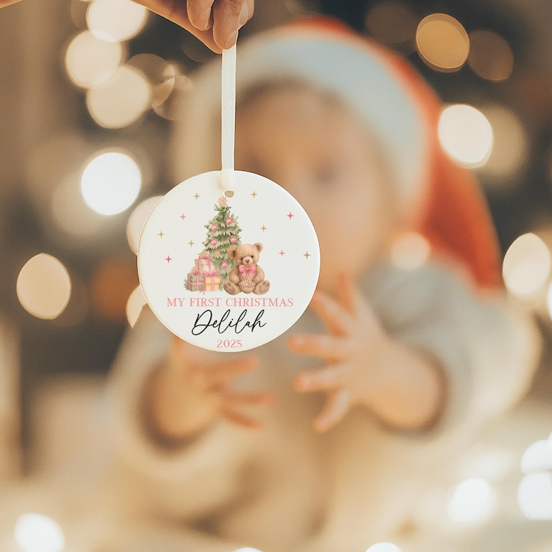 May include: A round Christmas ornament with a festive design. The ornament features a Christmas tree, gifts, and a teddy bear illustration in soft pink and brown hues. The text 'MY FIRST CHRISTMAS Delilah 2025' is displayed. A white ribbon is attached for hanging.