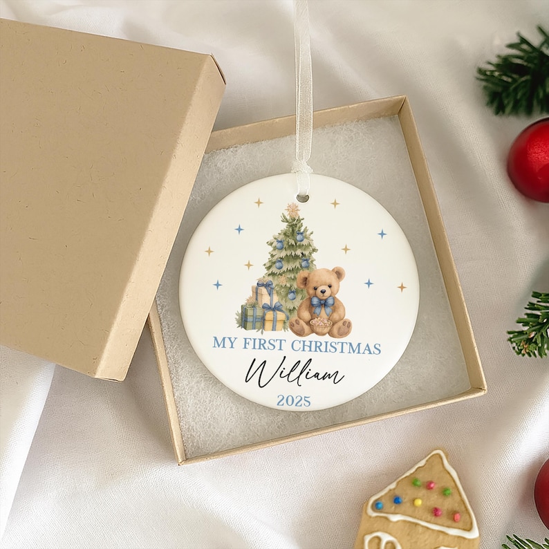 May include: A round Christmas ornament with a festive design. The ornament features a Christmas tree, a teddy bear with a blue bow, and gifts. The text 'MY FIRST CHRISTMAS' and the name 'William' with the year '2025' are displayed. The ornament is white with blue and gold accents and has a ribbon for hanging.