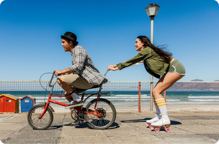 A split-screen with a line in the middle shows a man and a woman near the beach. On the left, which appears slightly blurry, the man is riding a bike. On the right side, which is sharp and clear, the woman is holding onto the man's bike wearing roller skates, highlighting the OIS feature of Galaxy A26 5G.