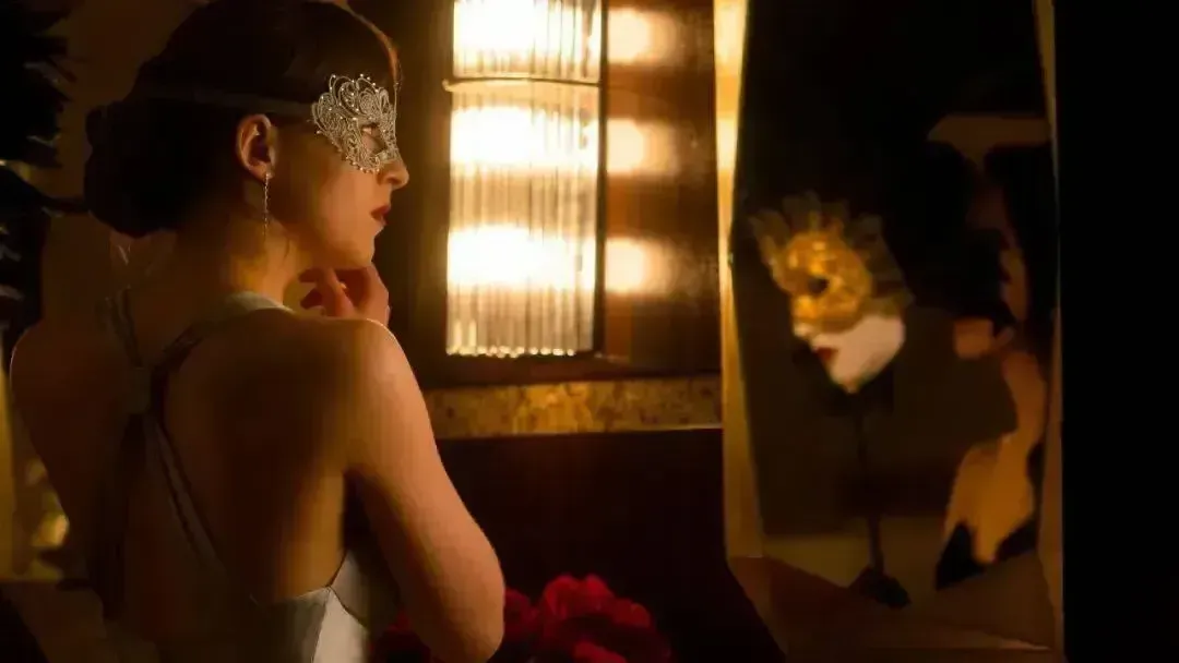 The picture shows the heroine of the movie 50 Shades of Grey wearing earrings in front of a mirror. She has a silver eye mask on her face. Other masks can be seen in the reflection of the mirror. The dim light illuminates a bouquet of roses.