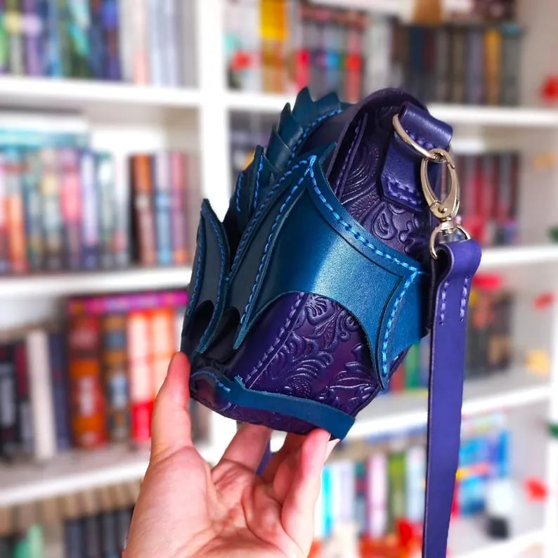 May include: A handcrafted, fantasy-themed crossbody bag in shades of blue and purple. The bag features a unique design with a dragon scale-like texture on the front, complemented by intricate embossed patterns. It has a long, adjustable strap and a silver-colored clasp.