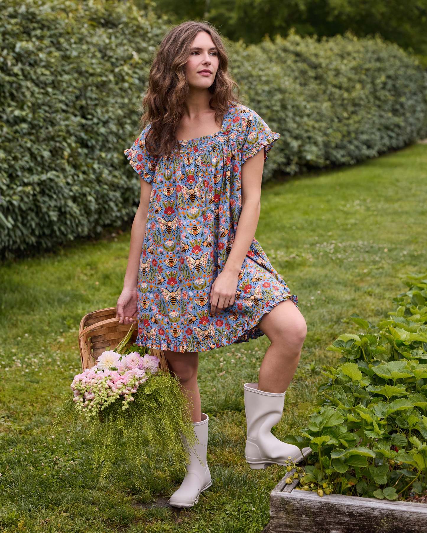 Person wearing the Delphinium Blue Bee Tapestry pintuck nightgown with short sleeves and a ruffled hem, standing beside a garden bed with flowers.