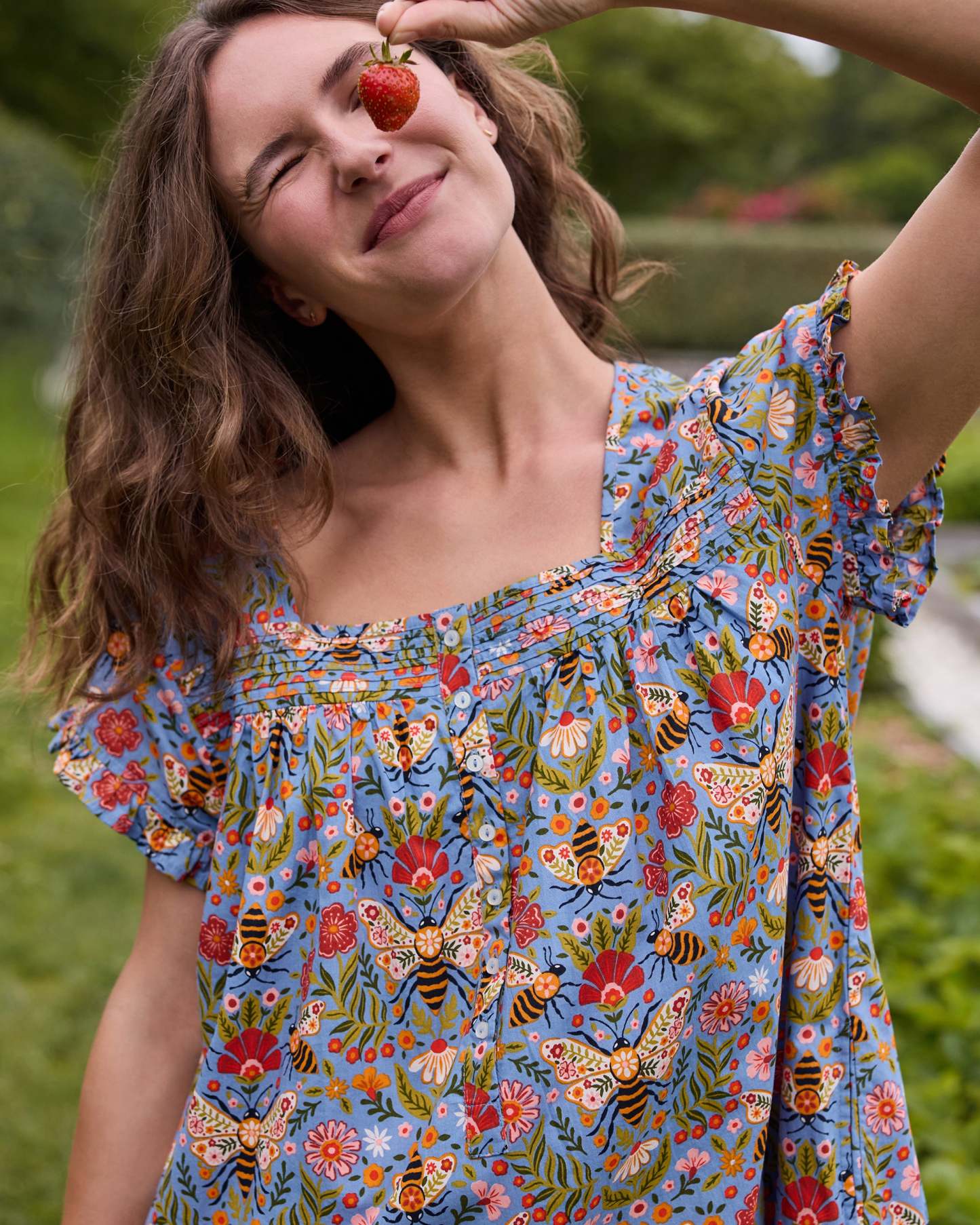 Close view of the Delphinium Blue Bee Tapestry nightgown's pintuck neckline, buttons, ruffled sleeves, and bee-and-floral print while the wearer holds a strawberry.