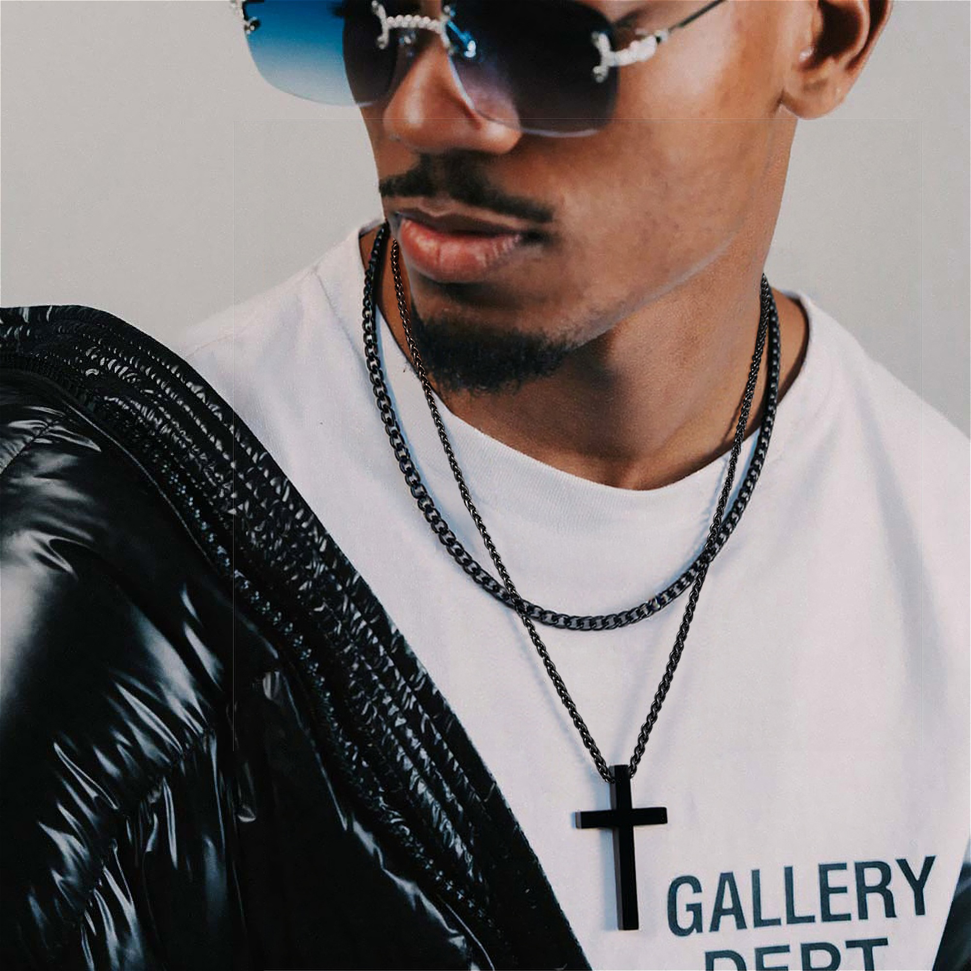 Bearded man in streetwear layering two black necklaces, one with a cross charm