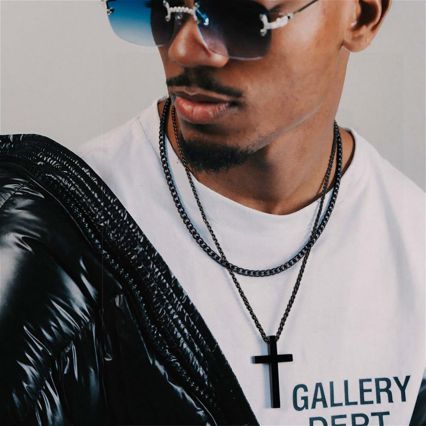 Bearded man in streetwear layering two black necklaces, one with a cross charm