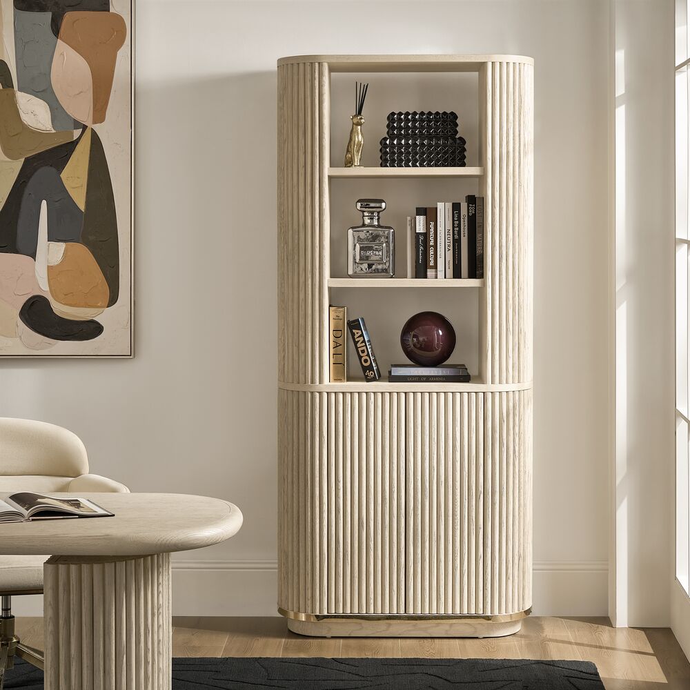 Modern tall fluted light wood arched bookcase with brass base styled with books and decorative accents next to an office desk.