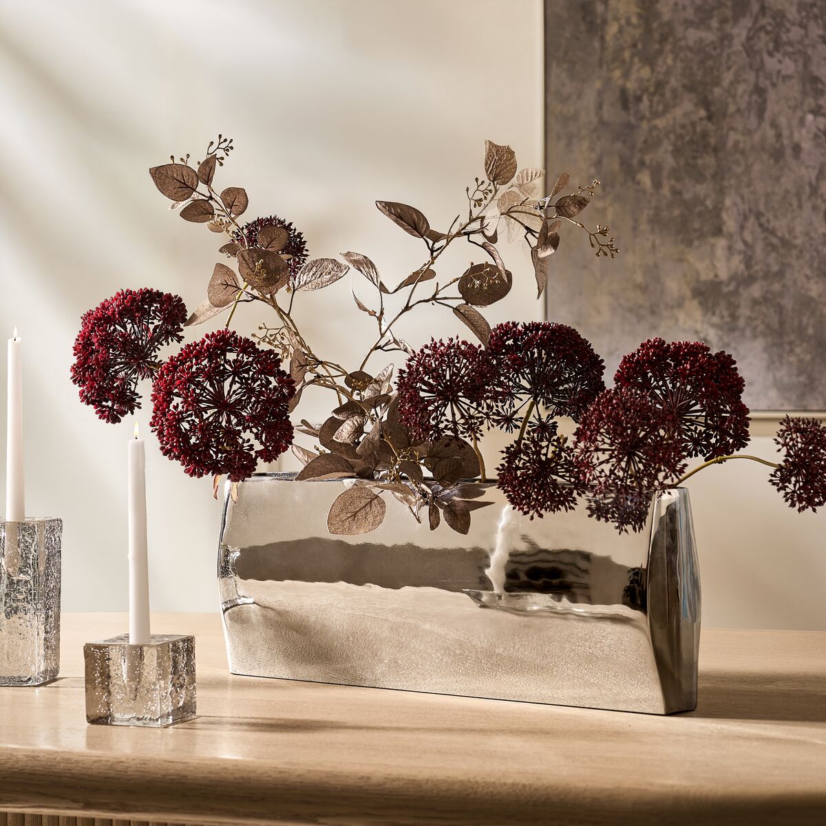 Modern mirrored trough vase filled with deep red floral stems alongside crystal candle holders on a tabletop display.