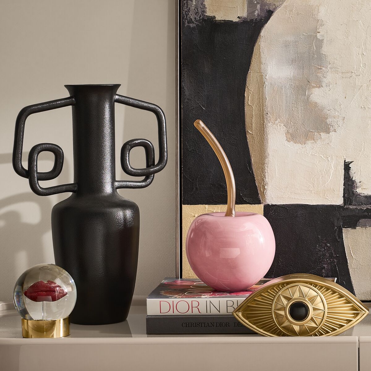 Eclectic home accents display including a sculpted black matte vase, glossy pink cherry object, golden evil eye decor, and designer books.