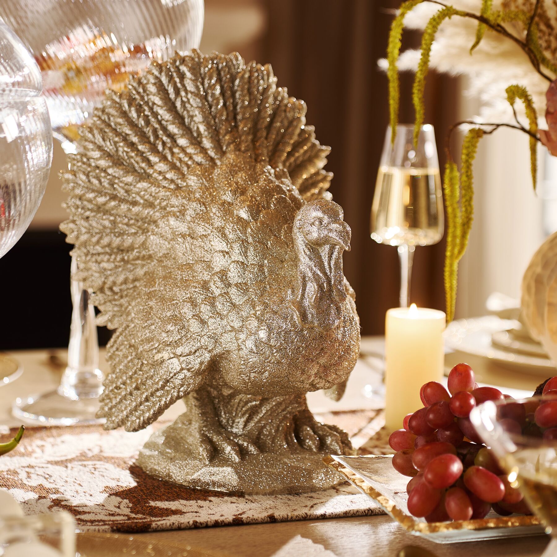 Elegant Thanksgiving dining table centerpiece featuring a textured metallic gold turkey sculpture, champagne glasses, glowing taper candles, and fresh grapes.