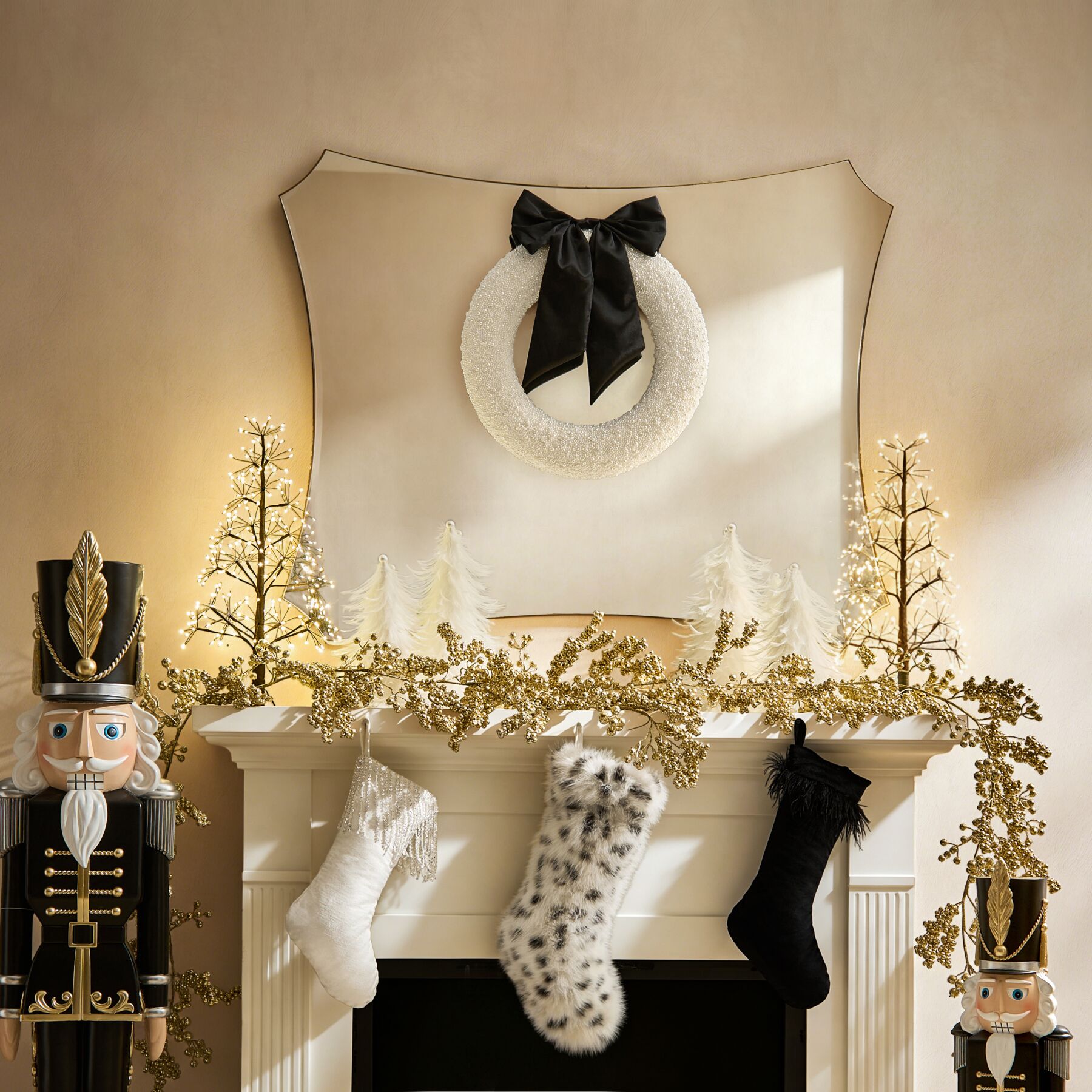 Festive Christmas mantel display styled with a black and gold nutcracker, illuminated mini trees, faux fur holiday stockings, and a white wreath with black bow.