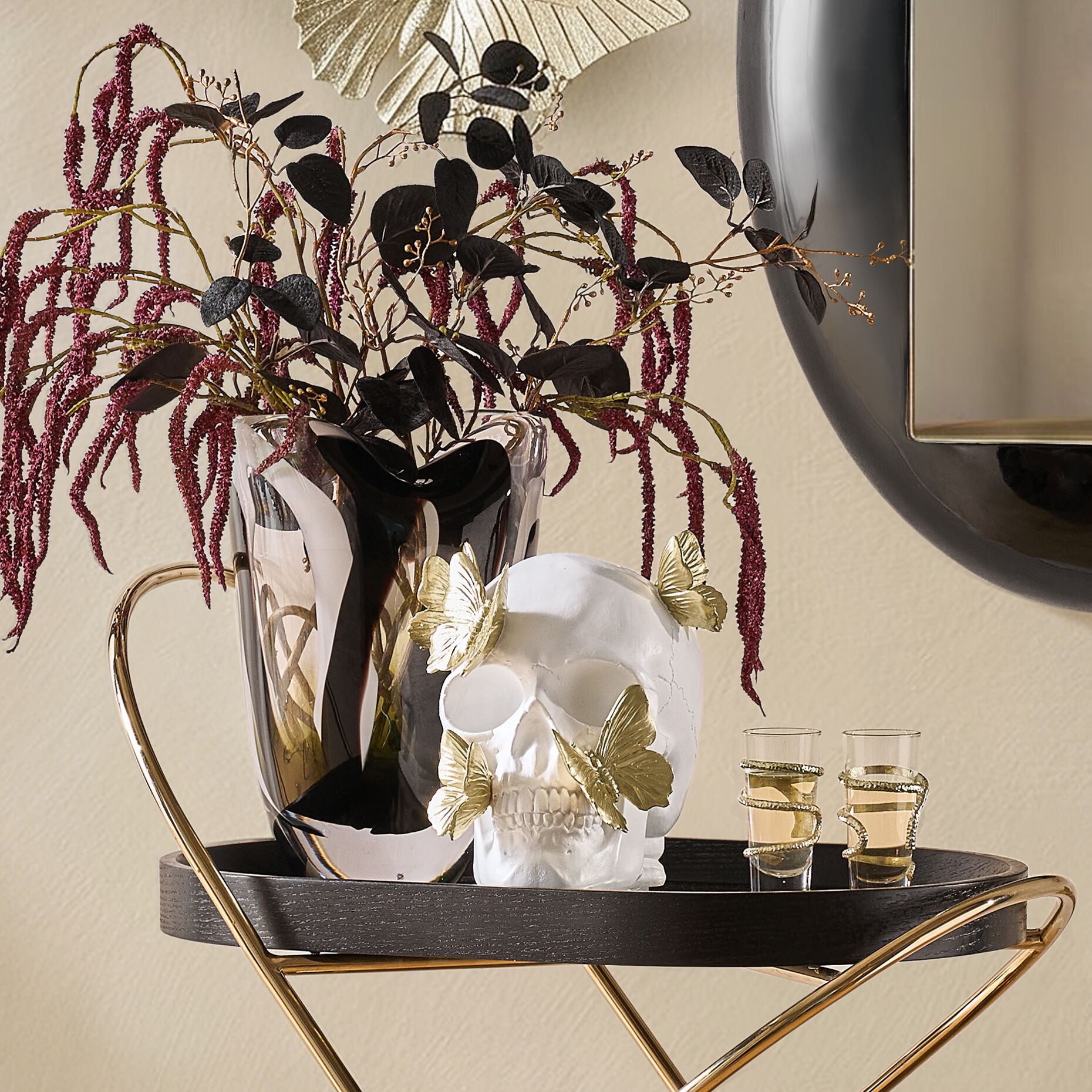 Chic Halloween bar cart vignette featuring a white skull sculpture with gold butterflies, dark floral arrangement, and cocktail glasses beside a modern mirror.