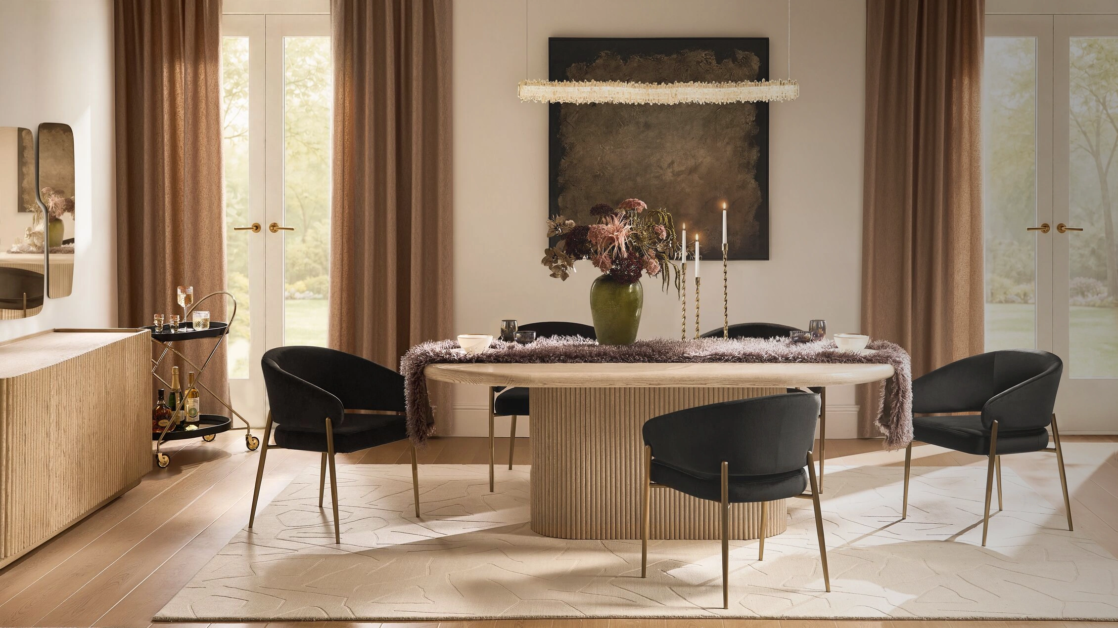 Modern dining room setup featuring a light wood fluted pedestal dining table, black velvet curved chairs with brass legs, crystal linear chandelier, fluted sideboard, and rolling bar cart.