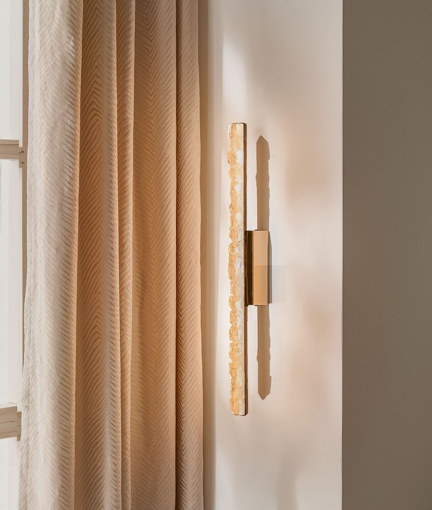 Minimalist vertical linear brass and alabaster wall sconce mounted on an off-white wall beside pleated drapery.