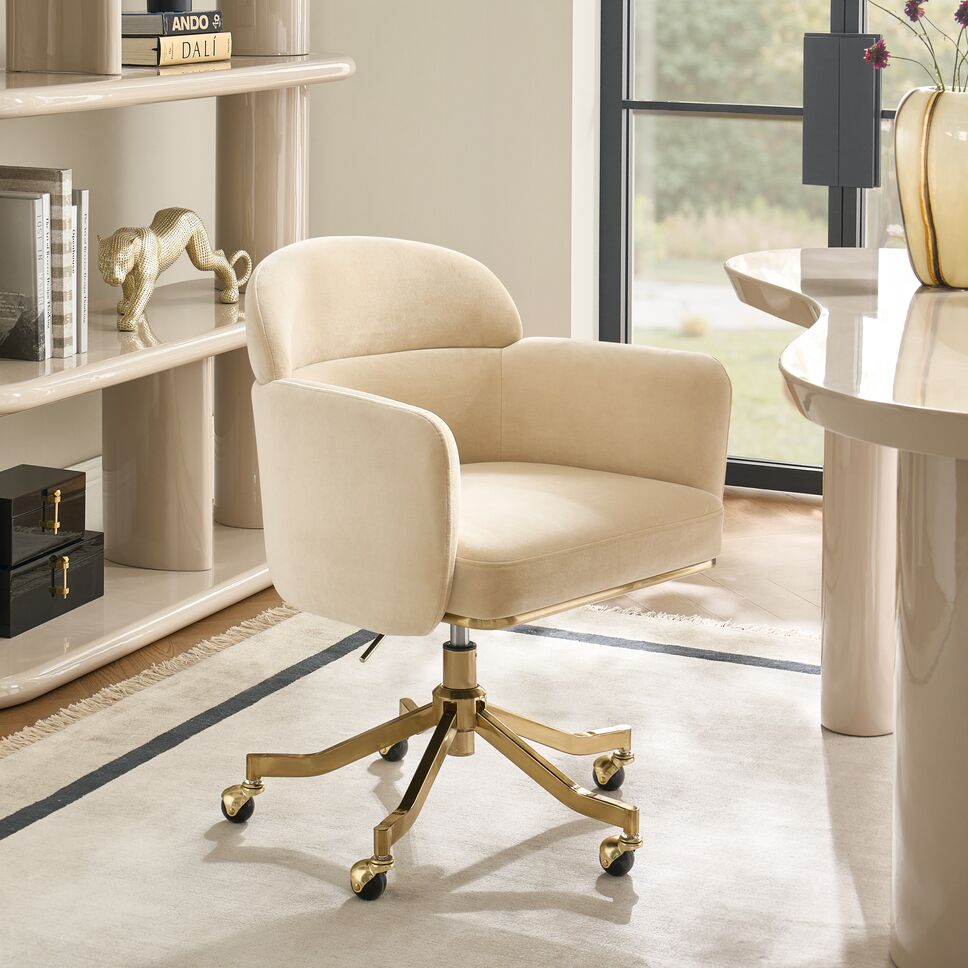 Ivory upholstered curved modern desk chair with brass swivel caster base in a sunlit home office studio.
