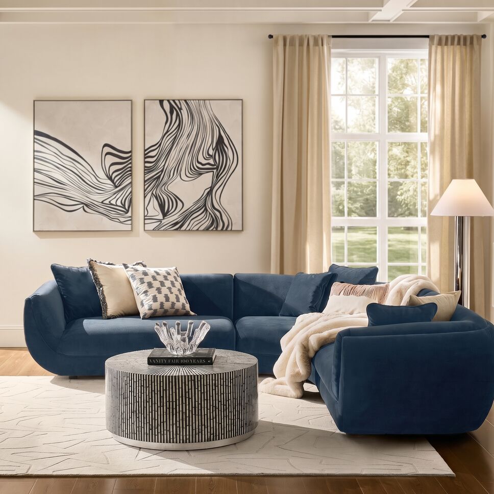 Modern curved deep blue velvet sectional sofa styled with patterned throw pillows, a round fluted cocktail table, and tall floor lamp in a sunlit living room.