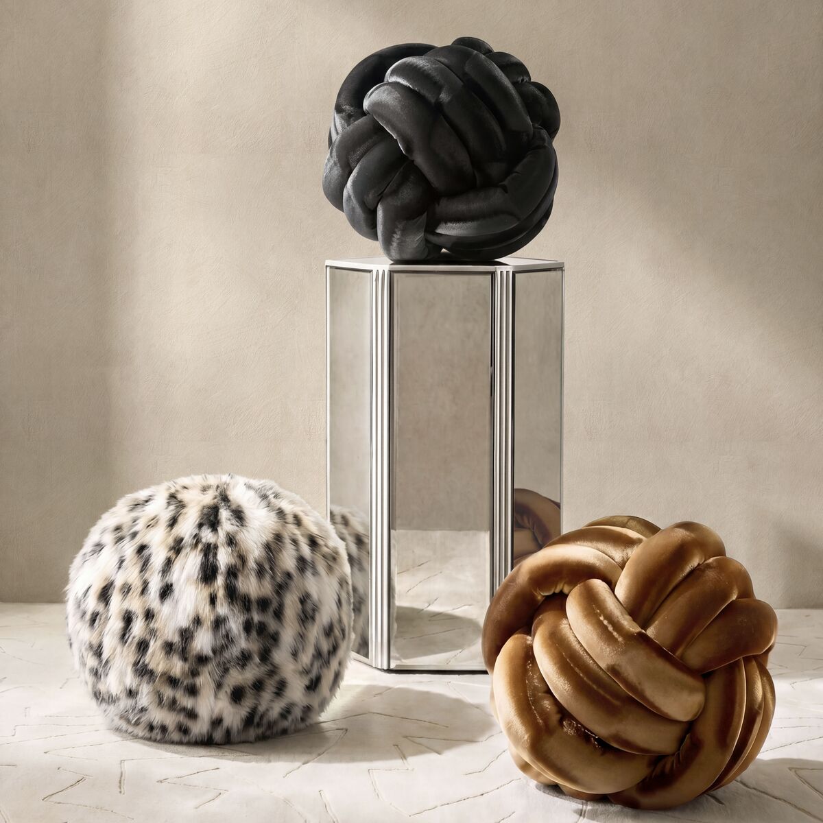 Decorative knot pillows in black leather, velvet camel, and leopard faux fur displayed on a mirrored pedestal.