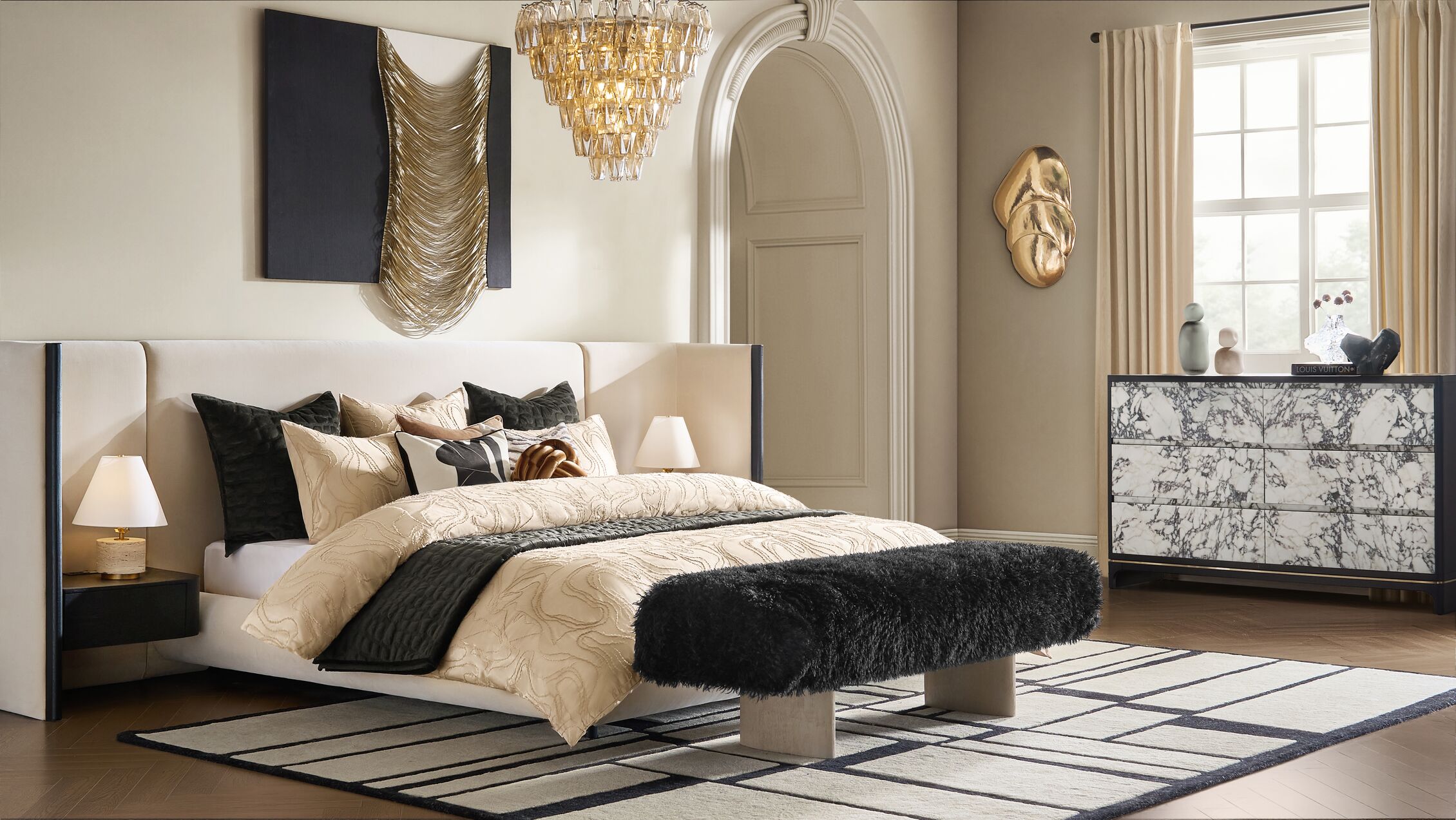 Spring Summer 2026 bedroom decor with neutral upholstered bed, layered pillows, amber glass chandelier, black faux fur bench, geometric area rug, and gold wall art.