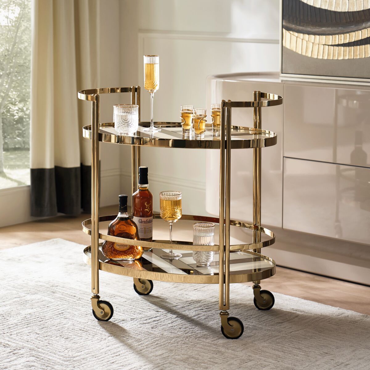 Oval two-tier brass bar cart with glass shelves, rolling casters, and barware accessories in a modern dining room.