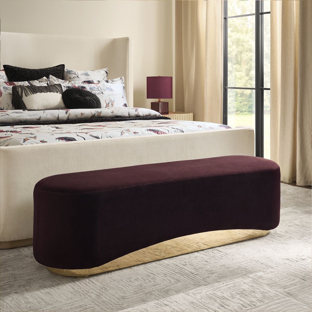 Modern curved deep burgundy velvet bedroom bench with a sleek gold base set at the foot of an upholstered bed.