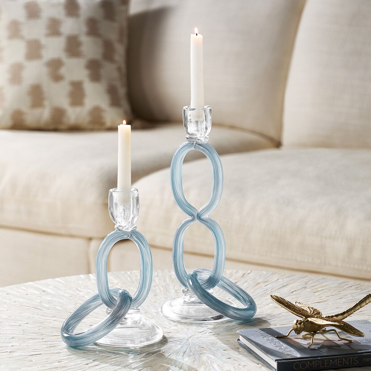 Modern light blue glass chain link taper candleholders holding white candles on a round living room coffee table.