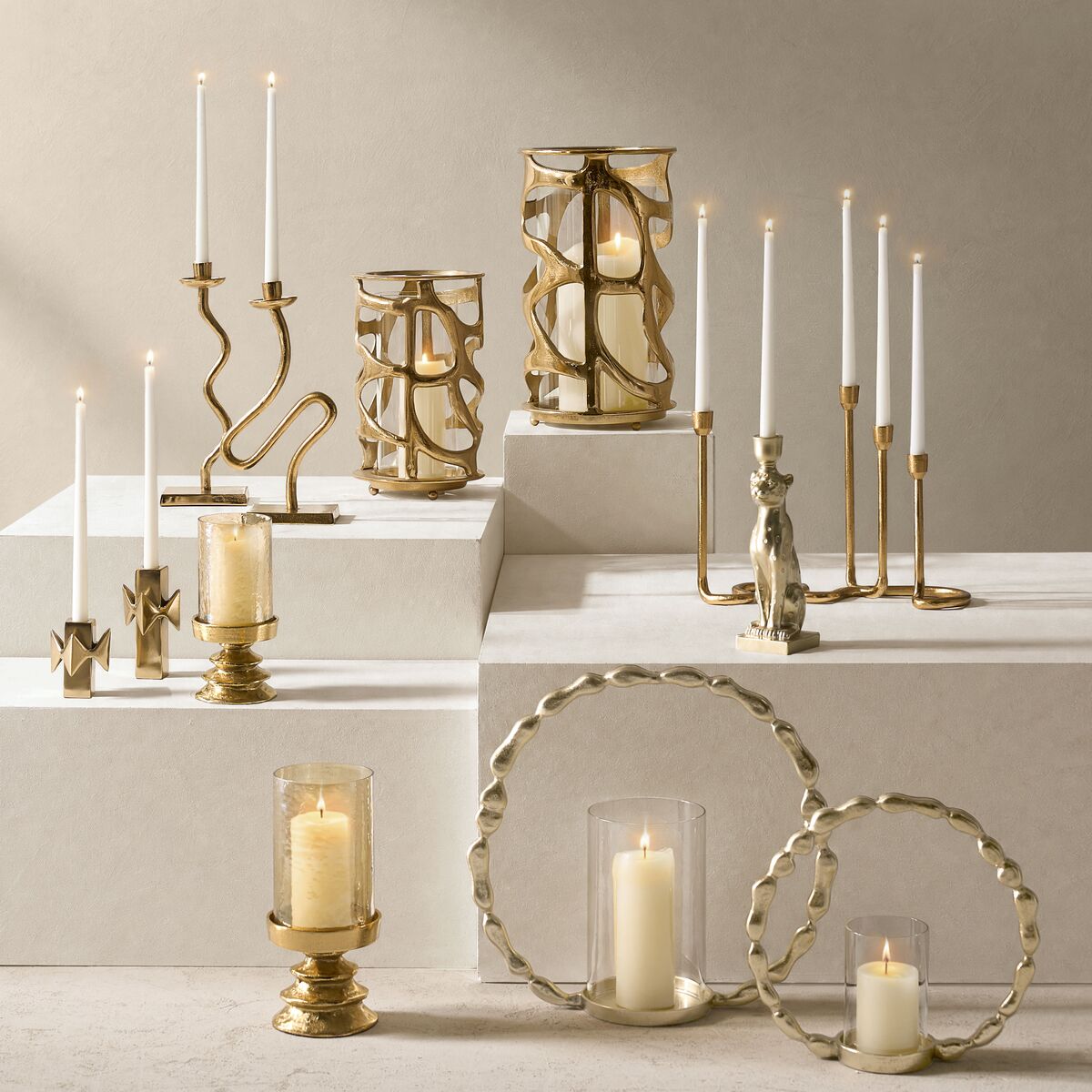 Assorted brass and gold candle holders including hurricane lanterns, taper candlestick holders, and circular brass stands with lit candles.